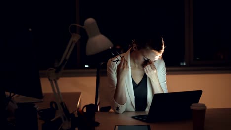 Exhausted-young-lady-is-working-in-office-late-at-night-using-laptop-then-taking-off-her-glasses-and-rubbing-face-relaxing.-Overwork-and-millennials-concept.