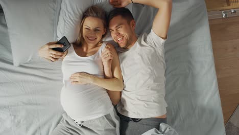 Amantes-de-la-joven-pareja-pasar-mañana-en-la-cama,-mujer-joven-embarazada-muestra-a-su-pareja-algo-en-un-Smartphone-de-pantalla-táctil,-Selfie-de-tomar-y-compartir-imágenes-en-redes-sociales.