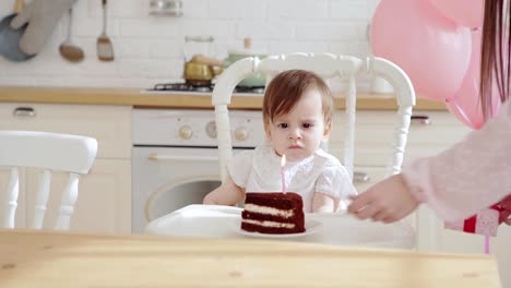 Liebevolle-Mutter-schenkt-Baby-Tochter-sitzen-im-Hochstuhl-ein-Stück-Geburtstagskuchen-mit-einer-brennenden-Kerze-auf-ihrer-ersten-Geburtstagsfeier