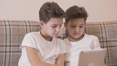 Close-up-faces-of-Caucasian-twin-brothers-playing-video-games-at-tablet-and-giving-high-five.-Siblings-having-fun-together-indoors.-Twins-resting-at-home.
