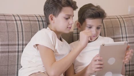 Close-up-portrait-of-two-Caucasian-twins-playing-video-games-at-tablet-and-giving-high-five.-Brothers-spending-free-time-together-indoors.-Siblings-resting-at-home.