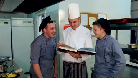 Joven-cocinero-a-aprendices-tener-discusión-positiva-con-chef-sosteniendo-el-libro-de-cocina