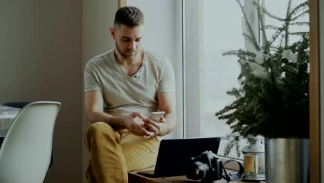 Attractive-young-man-browsing-smartphone-phone-sitting-on-windowsill-with-laptop-and-camera-in-cafe