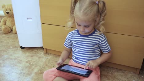 Baby-Mädchen-hält-eine-Tablette-und-sitzt-auf-dem-Boden-im-Zimmer.