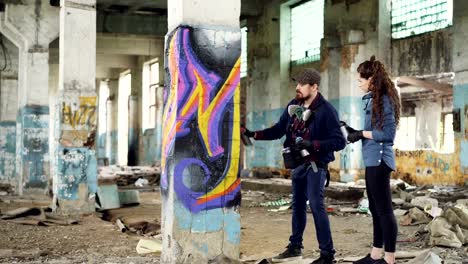 Experienced-graffiti-artist-is-teaching-his-friend-to-create-beautiful-images-with-spray-paint,-they-are-standing-together-in-adandoned-building-near-old-column-and-talking.