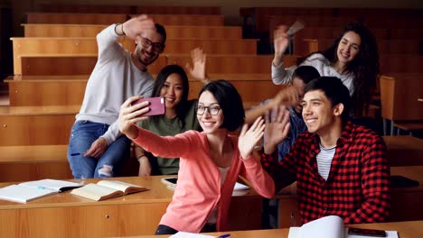 Freudige-Studenten-haben-Spaß-Aufnahme-video-im-Klassenzimmer-winkenden-Händen-und-lächelnd-an-Schreibtischen-im-Hörsaal-sitzen.-Selfie,-Smartphone-und-Bildung-Konzept.