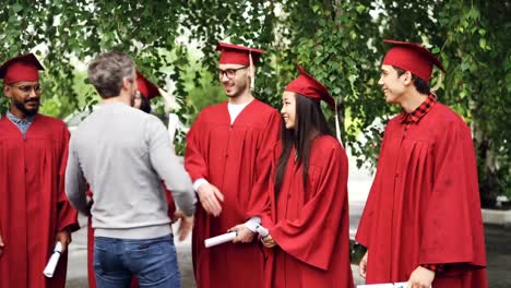 Bärtiger-Mann-stolz-Lehrer-ist-gratuliert-Absolventen-Händeschütteln-und-umarmt-sie-im-Freien-im-College-Campus,-während-Studenten-sprechen-und-hält-Diplome.