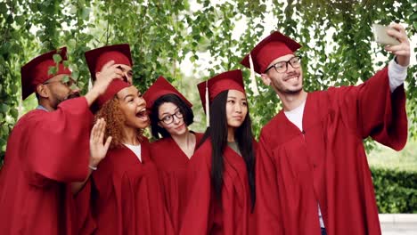 Excited-young-people-graduates-are-taking-selfie-with-smartphone,-boys-and-girls-are-posing,-waving-hands,-smiling-and-laughing.-Self-portrait-and-youth-concept.