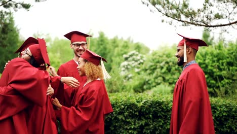 De-alegres-chicas-y-chicos-graduar-estudiantes-abrazar-y-estrechar-las-manos,-usar-sombreros-y-vestidos-de-graduación-día-al-aire-libre-disfrutando-de-lenta.