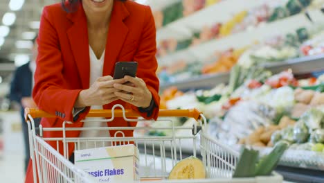 En-el-supermercado:-mujer-usa-Smartphone,-se-inclina-en-el-carrito-en-el-fresco-Produce-sección-de-la-tienda.-En-la-mujer-de-gran-centro-de-navegación-en-Internet-en-su-teléfono-móvil.-Cámara-lenta.