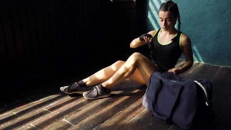 Panning-young-beautiful-woman-sitting-on-floor-and-wrapping-hands-with-black-boxing-wraps-in-club.