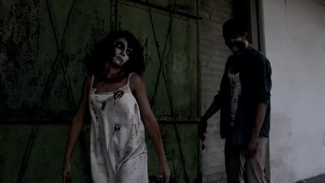 Two-zombies-are-walking-with-an-abandoned-house-on-the-background.-Brunette-girl-with-wounded-face-and-bloody-white-dress-and-wounded-male-zombie-are-walking-outdoors