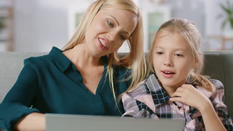 Beautiful-Young-Mom-and-Her-Cute-Little-Daugther-Use-Laptop-while-Sitting-on-a-Sofa-at-Home.-Family-Spending-Time-Together-Watching-Videos-and-Cartoons-on-Computer.