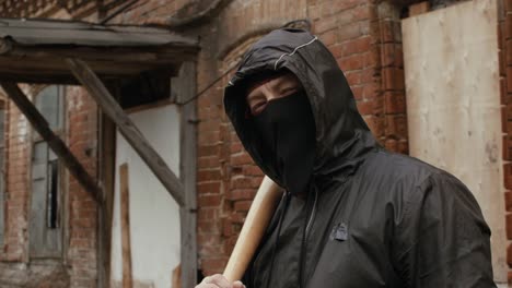 Street-hooligan-in-black-mask-with-baseball-bat-on-criminal-street-looking-to-camera