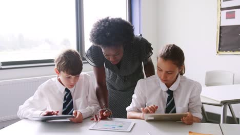 Profesor-de-secundaria-de-la-mujer-en-la-mesa-con-los-estudiantes-con-uniforme-con-tabletas-digitales-en-lección