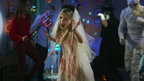 Halloween-Costume-Party:-Little-Girl-in-a-Bloody-White-Bride-Dress-Turns-Around-and-Makes-Scary-Faces.-In-the-Background-Group-of-Monsters-Dancing-and-Having-Fun-in-Decorated-Room-with-Disco-Lights