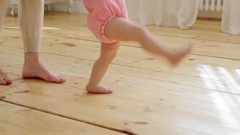 Tracking-shot-of-unrecognizable-cute-baby-girl-learning-to-walk-at-home-supported-by-her-loving-mother