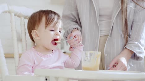 Madre-de-cuidado-irreconocible-alimentando-a-su-bebé-hija-puré-de-fruta-con-cuchara.-Chica-negándose-a-comer.-Mamá-limpiando-sus-manos-y-trona-con-tejido