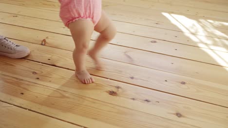 Tracking-shot-of-unrecognizable-caring-mother-supporting-her-adorable-baby-daughter-learning-to-walk-and-making-her-first-steps-on-hardwood-floor-at-home