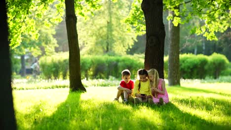 Foto-de-seguimiento-de-tres-niños,-dos-niñas-y-niño,-sentado-en-hierba-verde-bajo-el-árbol-en-el-parque-y-jugando-videojuegos-en-el-teléfono-inteligente