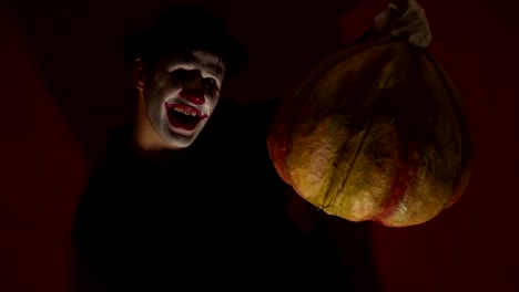 El-payaso-aterrador-tiene-una-máscara-de-calabaza-en-sus-manos.-Un-hombre-aterrador-con-maquillaje-de-payaso-sostiene-una-calabaza-para-Halloween.-Un-payaso-de-miedo-tiene-un-Jack-O-Lantern-en-sus-manos-con-ojos-luminosos-y-una-boca.