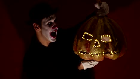 El-payaso-aterrador-tiene-una-máscara-de-calabaza-en-sus-manos.-Un-hombre-aterrador-con-maquillaje-de-payaso-sostiene-una-calabaza-para-Halloween.-Un-payaso-de-miedo-tiene-un-Jack-O-Lantern-en-sus-manos-con-ojos-luminosos-y-una-boca.