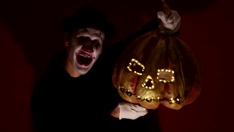 El-payaso-aterrador-tiene-una-máscara-de-calabaza-en-sus-manos.-Un-hombre-aterrador-con-maquillaje-de-payaso-sostiene-una-calabaza-para-Halloween.-Un-payaso-de-miedo-tiene-un-Jack-O-Lantern-en-sus-manos-con-ojos-luminosos-y-una-boca.