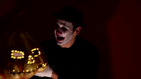 Scary-clown-holds-a-pumpkin-mask-in-his-hands.-A-scary-man-in-a-clown-makeup-holds-a-pumpkin-for-Halloween.-A-scary-clown-holds-a-Jack-O-Lantern-in-his-hands-with-luminous-eyes-and-a-mouth.