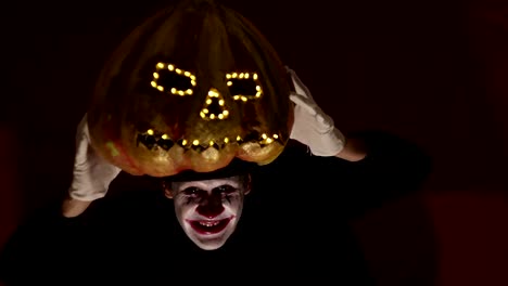 Scary-man-in-the-makeup-of-a-clown-holds-a-pumpkin-for-Halloween-and-puts-it-on-his-head.Terrible-clown-puts-on-a-mask-in-the-shape-of-a-pumpkin-on-his-head.-Terrible-clown-holds-in-his-hands-a-Jack-O-Lantern-with-luminous-eyes-and-a-mouth.