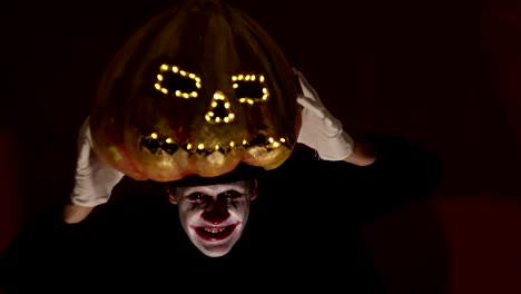 El-hombre-aterrador-en-la-composición-de-un-payaso-sostiene-una-calabaza-para-Halloween-y-se-la-pone-en-la-cabeza.-Un-payaso-terrible-se-pone-una-máscara-en-forma-de-calabaza-en-la-cabeza.-Terrible-payaso-sostiene-en-sus-manos-un-Jack-O-Lantern-con-ojos-