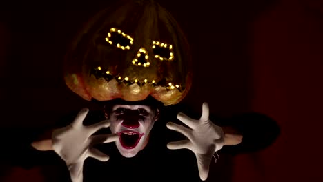 El-hombre-aterrador-en-la-composición-de-un-payaso-sostiene-una-calabaza-para-Halloween-y-se-la-pone-en-la-cabeza.-Un-payaso-terrible-se-pone-una-máscara-en-forma-de-calabaza-en-la-cabeza.-Terrible-payaso-sostiene-en-sus-manos-un-Jack-O-Lantern-con-ojos-