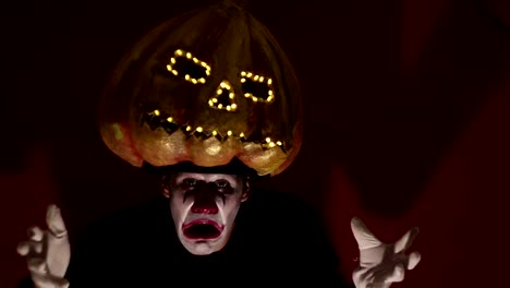 El-hombre-aterrador-en-la-composición-de-un-payaso-sostiene-una-calabaza-para-Halloween-y-se-la-pone-en-la-cabeza.-Un-payaso-terrible-se-pone-una-máscara-en-forma-de-calabaza-en-la-cabeza.-Terrible-payaso-sostiene-en-sus-manos-un-Jack-O-Lantern-con-ojos-