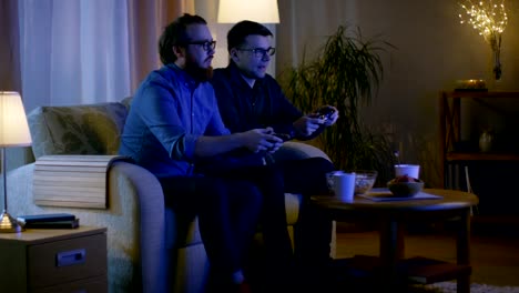 In-the-Evening-sitzen-zwei-Freunde-auf-einem-Sofa-im-Wohnzimmer-und-spielen-wettbewerbsfähige-Videospiele.