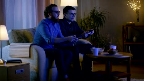 En-la-noche-dos-amigos-están-sentados-en-un-sofá-en-la-sala-de-estar-y-jugando-juegos-de-video-competitivos.