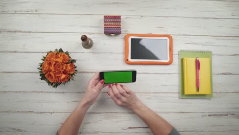 Frau-nimmt-eine-Smartphone-von-einem-weißen-hölzernen-Schreibtisch-und-tippen-mit-ca.-in-der-Nähe-sind-Blumen-und-eine-orange-digital-Tablette.-Chroma-Key-Green-Screen.-Ansicht-von-oben.-Hände,-Nahaufnahme