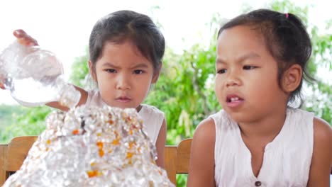 Two-asian-little-girls-do-the-baking-soda-and-vinegar-volcano-experiment-at-the-table-in-their-house,-slow-motion-in-100-fps