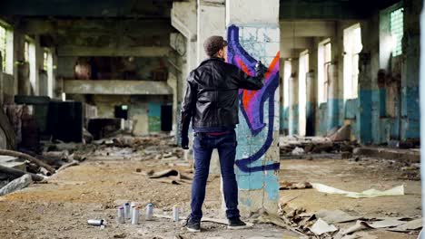 Back-view-of-graffiti-painter-creating-beautiful-image-with-aerosol-paint-inside-abandoned-building.-Artist-is-wearing-blue-jeans,-black-leather-jacket,-cap-and-gloves.