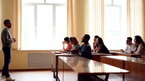 Freundliche-Lehrer-ist-im-Gespräch-mit-Studenten,-intelligente-Mischlinge-Mann-Hand-heben-und-sitzen-am-Tisch-im-Hörsaal-zu-sprechen,-während-seine-Mitschüler-schreiben.