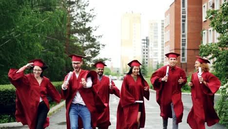 Emocionados-a-los-estudiantes-graduados-que-corre-a-lo-largo-de-la-carretera-en-el-campus-con-diplomas-usando-sombreros-y-vestidos-de-ropa-de-la-graduación.-Pequeña-lluvia-es-visible