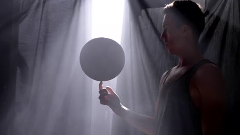 Basketball-player-spinning-ball-on-his-one-finger,-doing-tricks-in-misty-room-with-floodlight-in-background