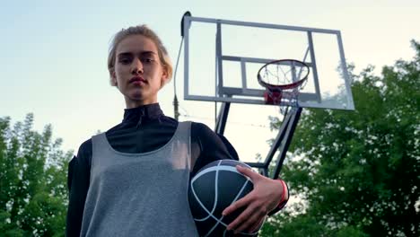 Beautiful-young-female-basketball-player-holding-ball-and-looking-at-camera,-standing-in-park,-low-angle