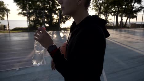 Down-up-footage-of-a-young-sporty-girl-basketball-player-have-a-rest-after-training-on-the-local-outdoors-court,-drinking-a-water-from-a-plastic-bottle