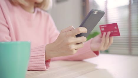 Beautiful-Asian-woman-using-smartphone-buying-online-shopping-by-credit-card-while-wear-sweater-sitting-on-desk-in-living-room-at-home.-Lifestyle-woman-at-home-concept.