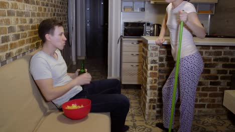 Man,-football-fan-is-drinking-beer-and-chips-in-front-of-TV,-the-wife-makes-cleaning