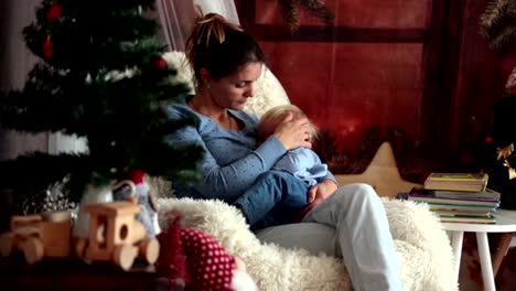 Mother-breastfeeding-her-toddler-son-sitting-in-cozy-armchair-near-Christmas-tree,-wintertime