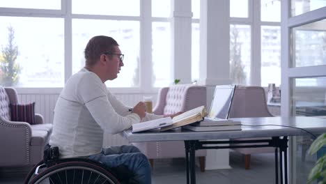 moderne-Technologien-in-der-Bildung,-Student-aus-Online-Unterricht-und-Notizen-im-Notizbuch-am-Tisch-im-Café-sitzen-lernen-Behinderte-im-Rollstuhl-mit-Laptop-arbeiten