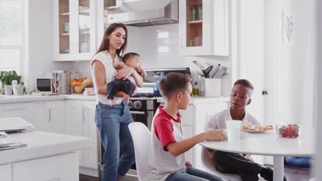 Madre-sosteniendo-bebé-se-encuentra-en-la-cocina-hablando-con-su-hijo-y-su-amigo,-más-de-una-fecha-de-juego