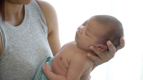 4K-Medium-close-up-dolly-shot-of-young-adult-asian-woman-mother-standing-near-window-in-home-bedroom-and-carrying-her-sleeping-nap-newborn-baby-girl-daughter.-Mother-day-and-baby-health-care-concept.