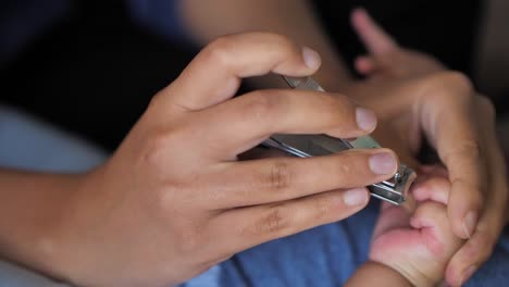 A-close-up-of-a-mixed-race-African-American-mother-clipping-one-of-her-baby's-fingernails-as-he-lays-on-his-back-on-the-bed-with-focus-on-their-hands-and-the-clipper-and-baby's-body-out-of-focus.