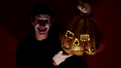 El-payaso-aterrador-tiene-una-máscara-de-calabaza-en-sus-manos.-Un-hombre-aterrador-con-maquillaje-de-payaso-sostiene-una-calabaza-para-Halloween.-Un-payaso-de-miedo-tiene-un-Jack-O-Lantern-en-sus-manos-con-ojos-luminosos-y-una-boca.